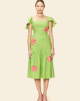 A woman faces forward in The Prana Bloom Linen Dress, a guava green cotton-linen midi featuring short ruffled sleeves and pink flower appliqués, paired with pink pointed shoes against a neutral background.