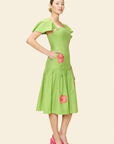 A woman stands in profile against a beige background wearing The Prana Bloom Linen Dress in guava green, featuring ruffled sleeves and hand-embroidered hibiscus, styled with bright pink heels.