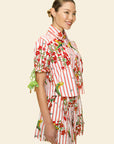 A woman smiles in The Maya Striped Cotton Shirt, featuring red vertical stripes with colorful floral and bird motifs, paired with matching shorts against a plain beige background.