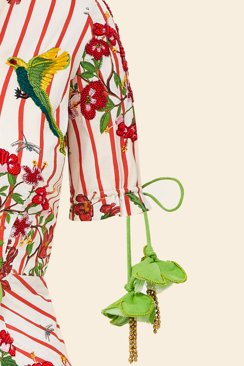 A close-up of The Maya Striped Cotton Dress shows red stripes, colorful floral and bird embroidery, plus green fabric flower accents with beaded tassels hanging from a green tie at the sleeve.