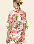 A woman with blonde hair in a low bun is seen from the back wearing The Maya Striped Cotton Shirt featuring red stripes and green parrot and floral prints, against a plain beige background.