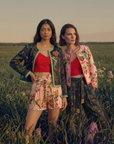 Two women stand confidently in a grassy field at sunset, surrounded by purple wildflowers. They wear matching jackets, red tops, and The Maya Striped Cotton Short.