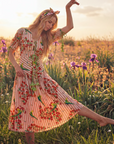 A woman with long blond hair poses in a sunlit meadow wearing The Maya Striped Embroidered Handkerchief as a headscarf and a floral, bird-print dress. Surrounded by purple irises, she strikes a graceful pose with one arm raised.
