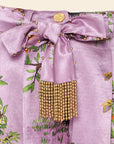 Close-up of The Lilac Shanti Tiger Pleated Shorts: high-waisted designer lavender satin with a vibrant floral print, accented by a tied bow with gold beads, tassel, and a gold button at the waistband.