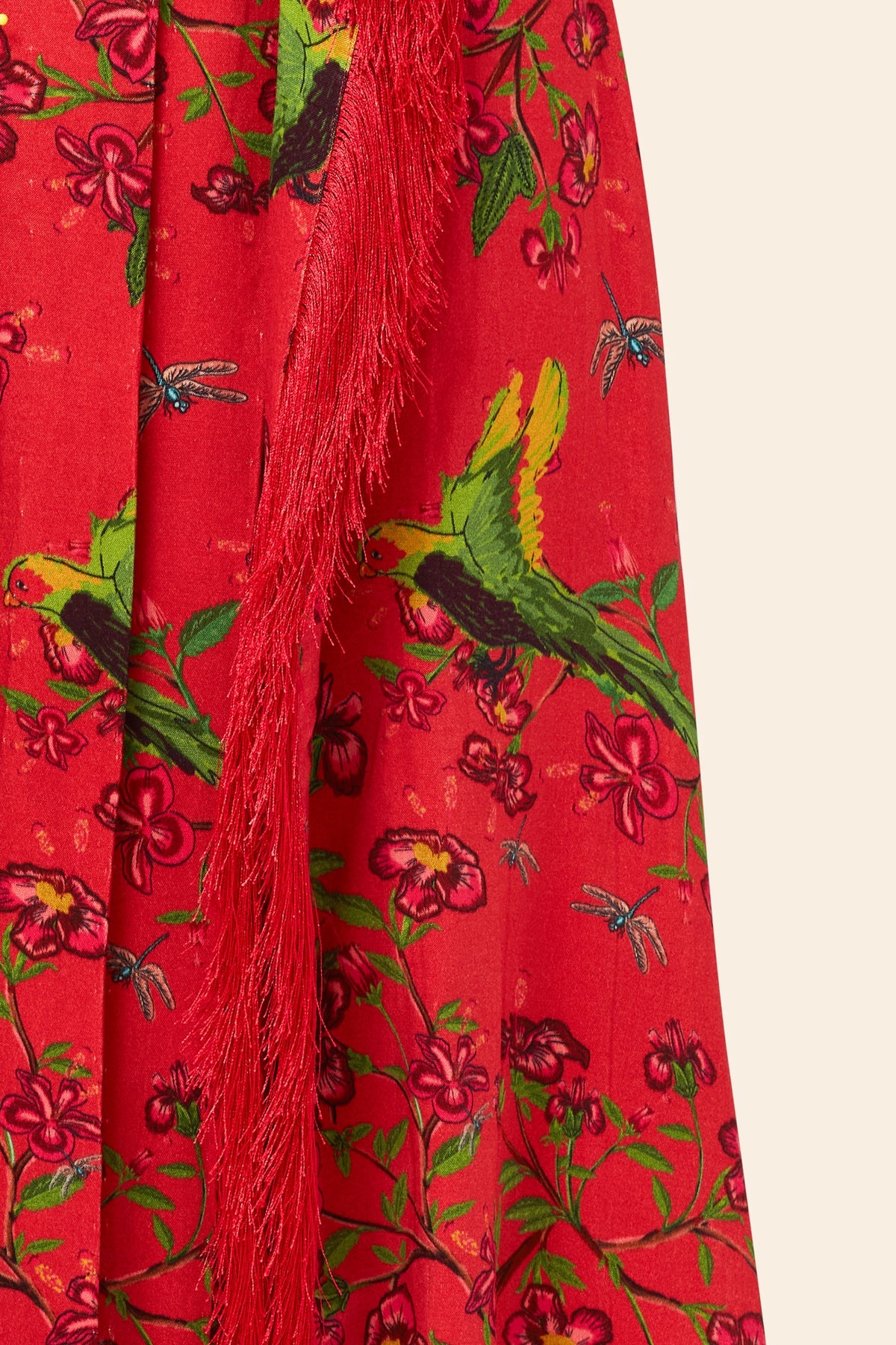 The Maya Fringe Skirt features bright red fabric with green parrots, pink flowers, blue dragonflies, a vertical red fringe trim, and subtle Aari embroidery for added texture.