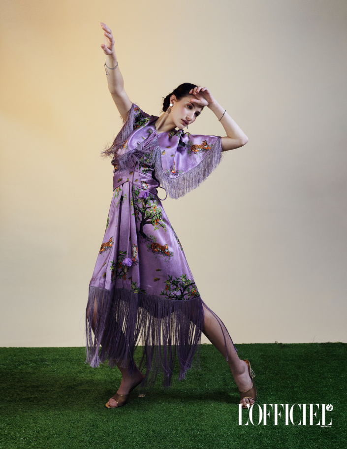 Woman in an Ashaki lilac dress with fringe standing on grass with a gradient background.