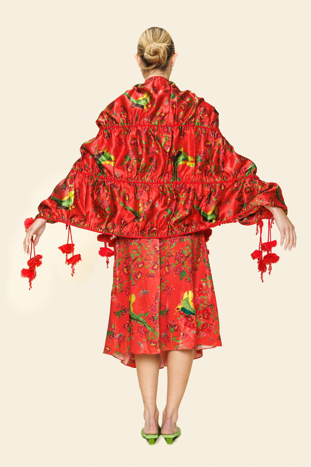 Woman wearing red floral skirt with fringe, red floral bra top and floral scarf with tassels and green parakeet print shown from the back
