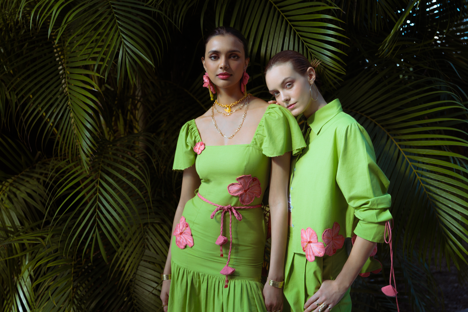 Two women in bright green outfits with pink floral details standing against a dark green leafy background.