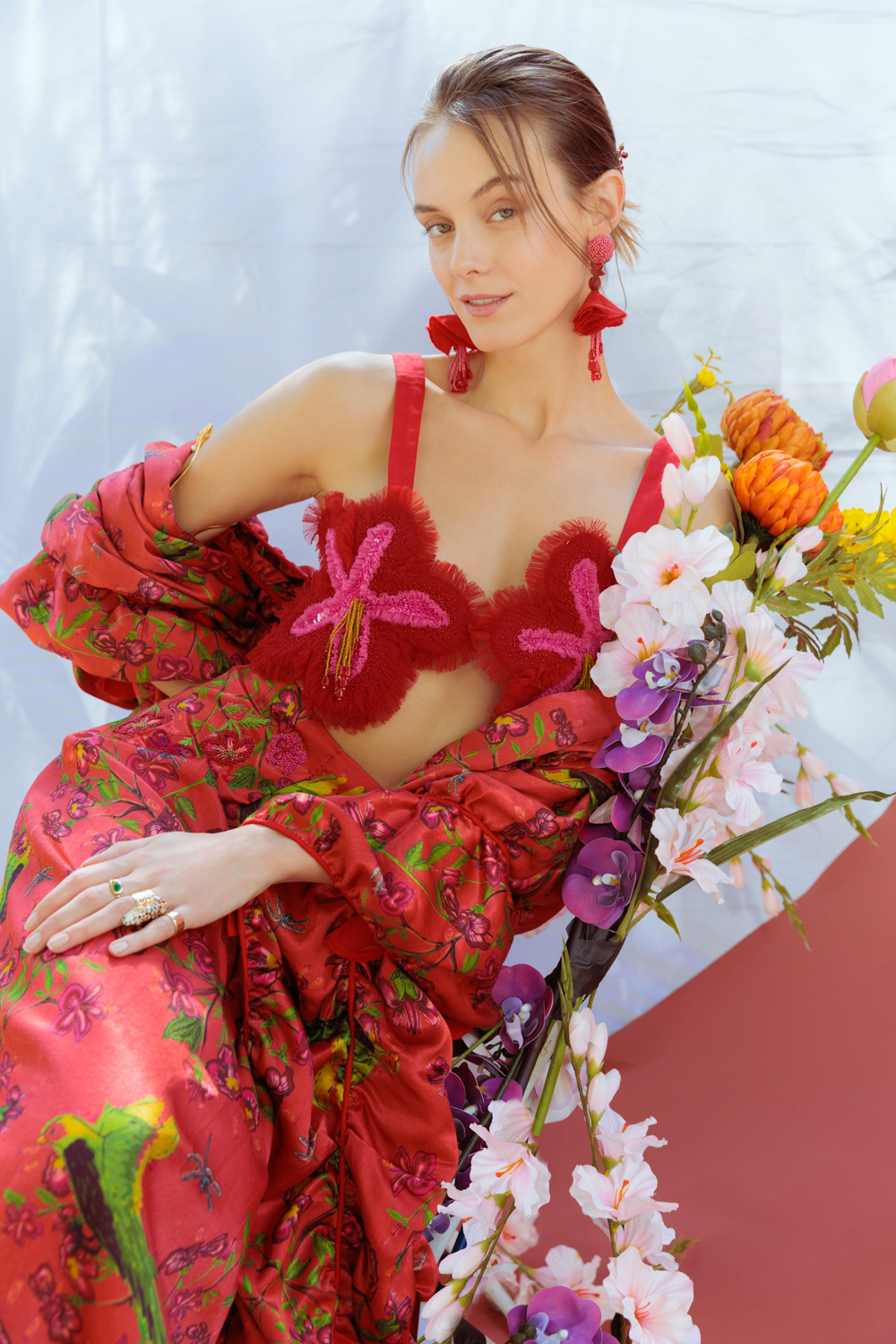 Woman in a vibrant red and pink outfit with floral elements, sitting next to a bouquet of flowers against a light blue background.