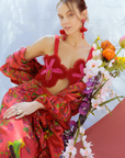 Woman in a vibrant red and pink outfit with floral elements, sitting next to a bouquet of flowers against a light blue background.