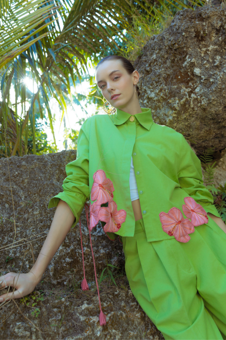 a woman in a bright green outfit with pink floral accessories standing among tropical plants.
