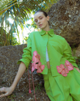 a woman in a bright green outfit with pink floral accessories standing among tropical plants.