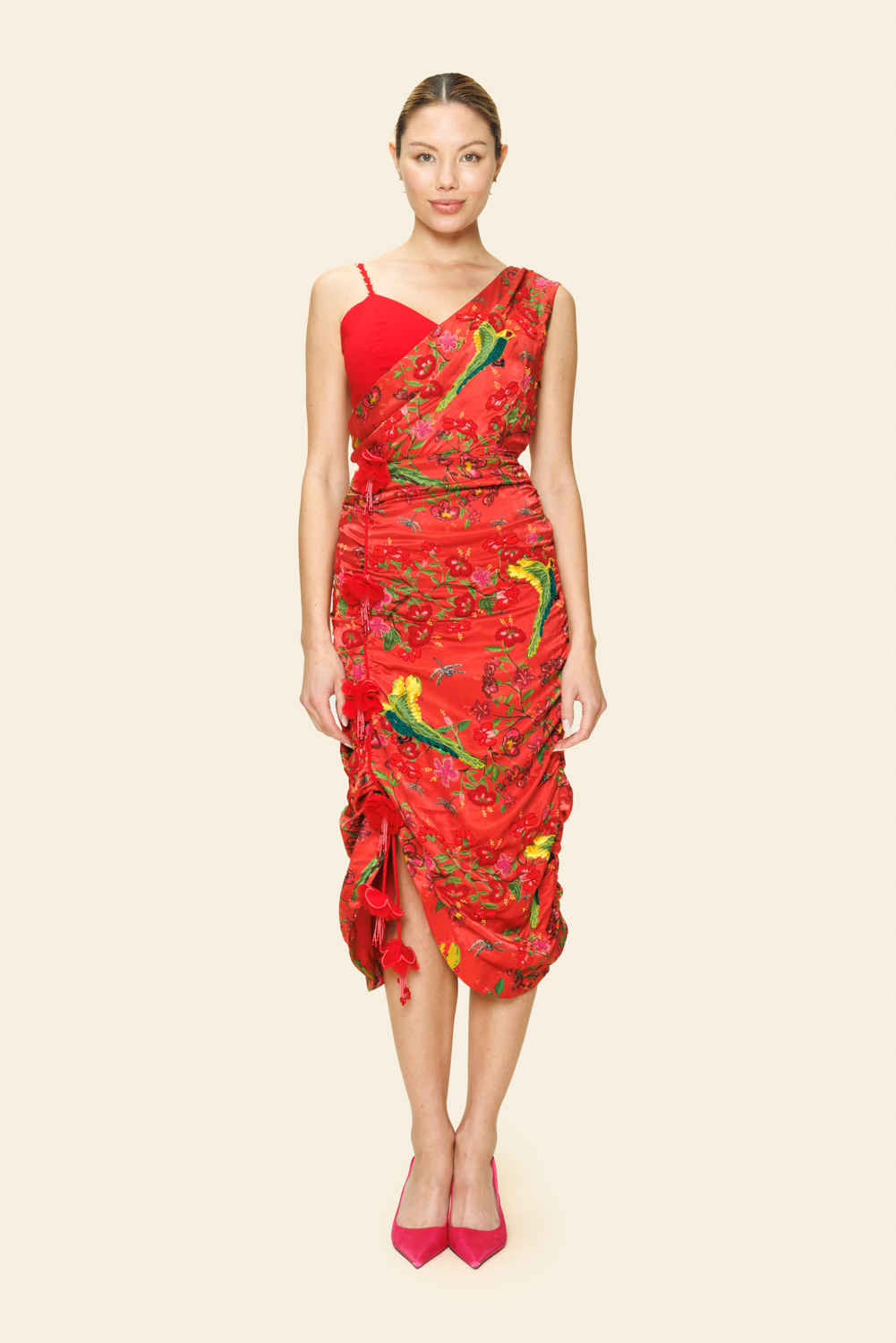 A model wearing a red one-shoulder draped midi dress with a floral pattern and beaded detail on the asymmetrical hem.