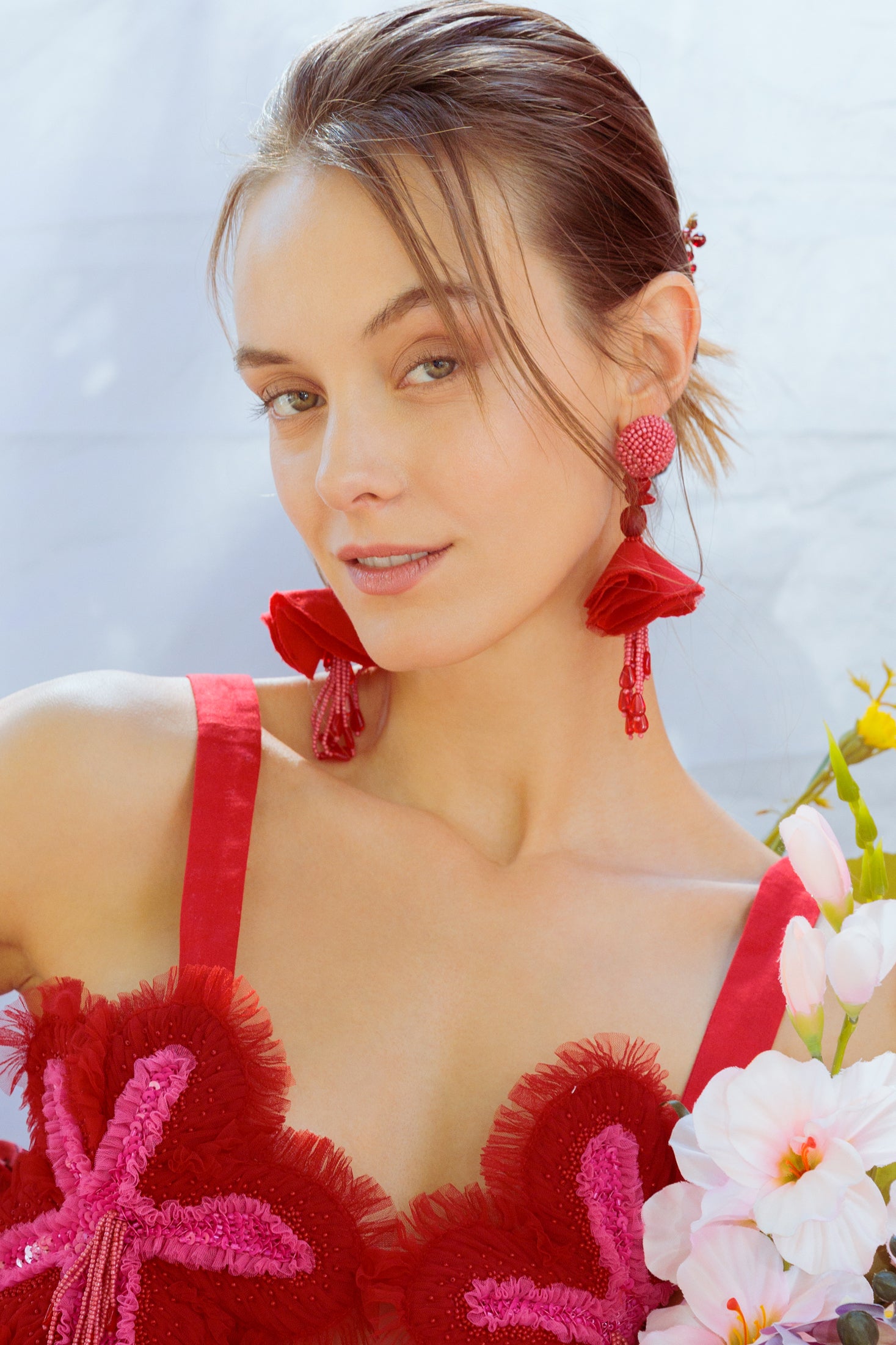 Woman wearing a top with floral details and red earrings, holding flowers against a light background 