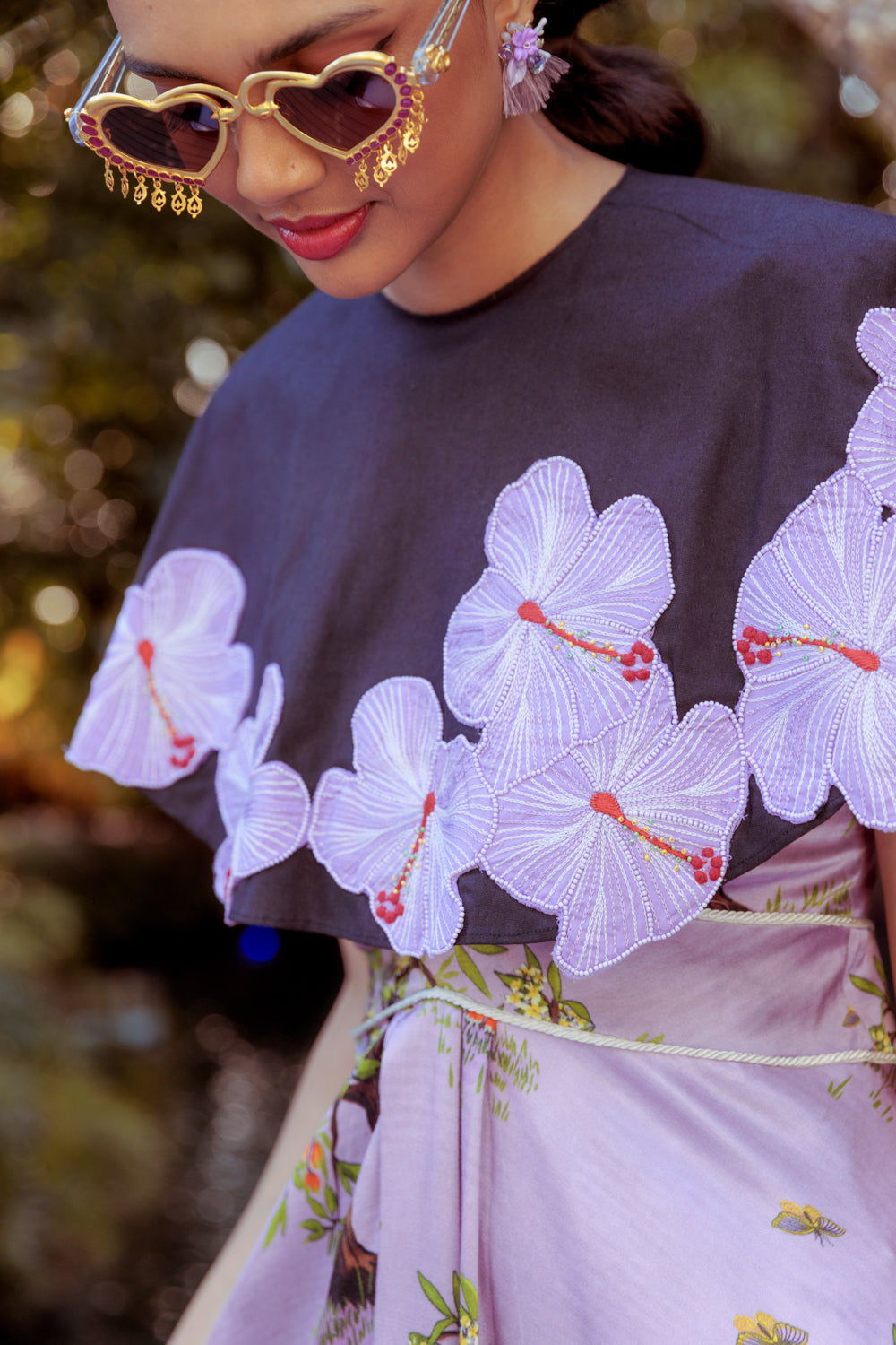 Person wearing a black cape top with floral embroidery over a pink dress against a blurred natural background.