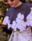 Person wearing a black cape top with floral embroidery over a pink dress against a blurred natural background.