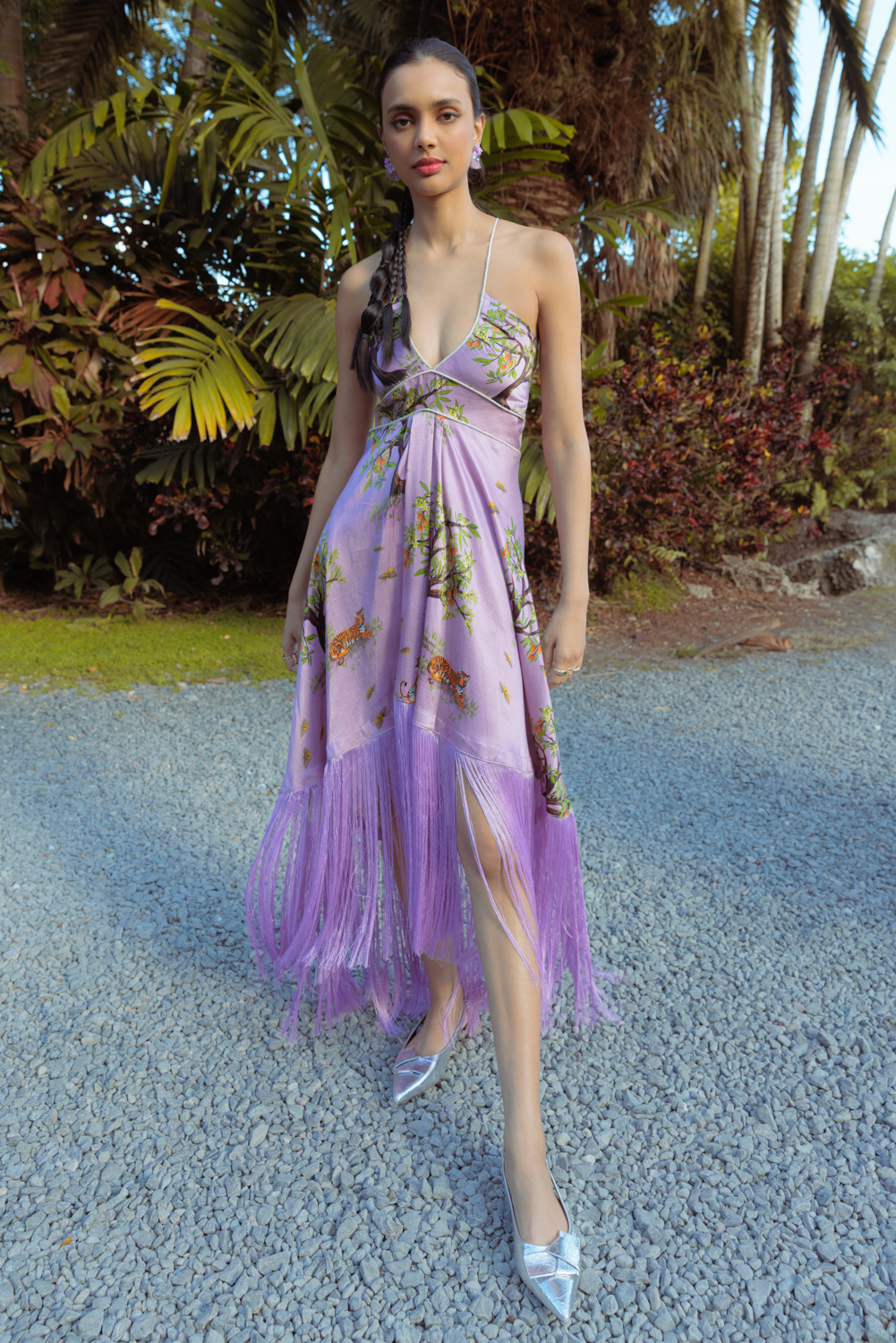 A woman wearing a purple midi dress with a tiger print, featuring fringe detailing on the skirt and adjustable rope straps. The dress has a pleated empire waist and is styled with silver heels.