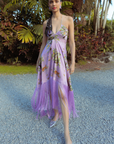 A woman wearing a purple midi dress with a tiger print, featuring fringe detailing on the skirt and adjustable rope straps. The dress has a pleated empire waist and is styled with silver heels.