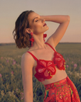 A woman with short brown hair stands in a grassy field at sunset, eyes closed and head tilted back. She wears a red floral bralette, a red skirt with colorful flowers, and The Maya Red Beaded Earring.