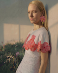 A woman with long blonde hair leans against a wall, wearing the Adhara White Cotton Sleeveless Top as soft sunlight casts shadows and blurred greenery appears in the background.